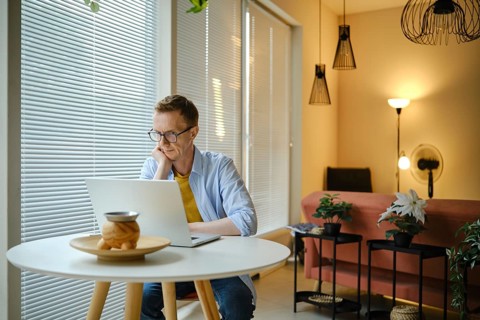 A man sits at a round table and watching sports on laptop, in a bright and airy living room.