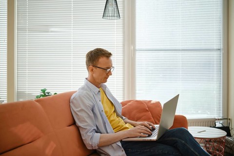 A retired man sits comfortably on a couch and writes his memoirs on laptop in a well-lit living