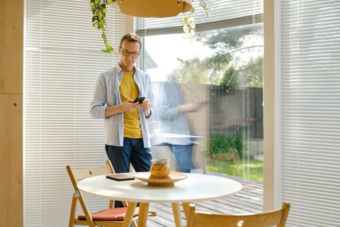 A middle-aged man stands in a bright, modern kitchen checking weather forecast at his smartphone.
