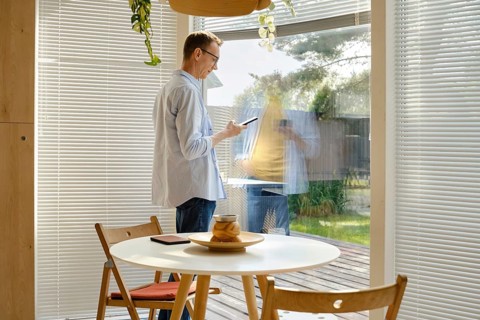 A man stands inside a stylish room, using his smartphone while looking out through a large glass