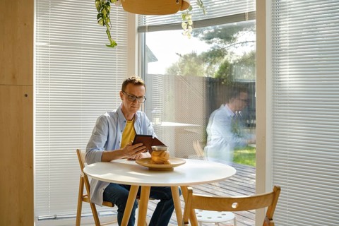 A man sits at a round wooden table in a modern kitchen, focused on his e-book. Sunlight filters