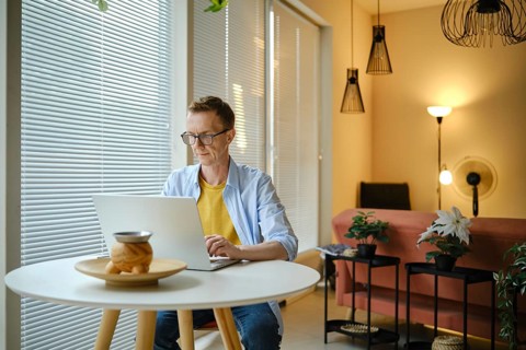 A freelancer is typing on a laptop at a round table in a modern home workspace. Natural light pours