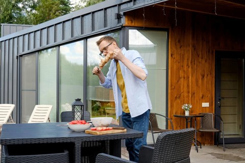A man is enjoying a summer barbecue at a stylish wooden house. He is focused on grilling meat,