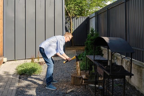A middle-aged man is chopping a piece of wood in a well-maintained backyard garden. He is dressed