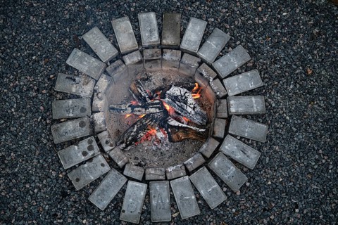 Bright flames dance among burning logs in a circular stone fire pit during early evening. The warm