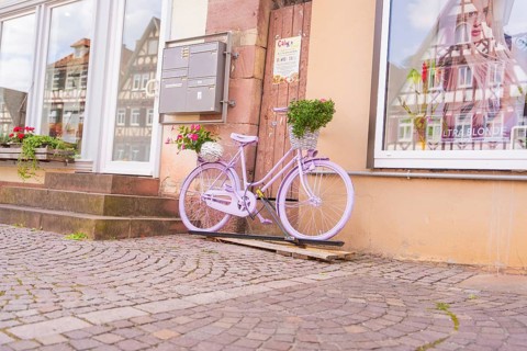 A purple bicycle with a basket of flowers in front of a building with half-timbered houses, small