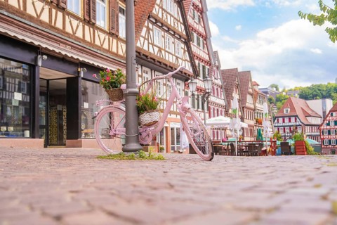 Pink bicycle with a basket of flowers stands on a cobbled street in front of half-timbered houses,
