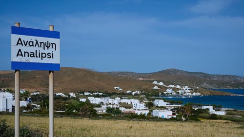 A town sign in front of a coastal settlement with whitewashed houses and blue sky, Analipsi,