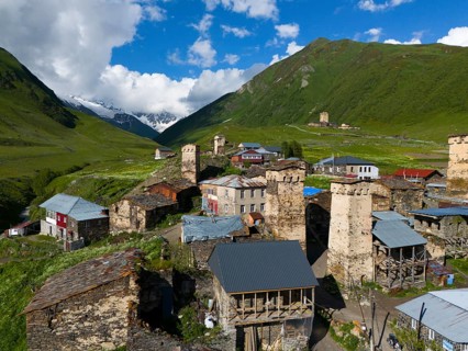 Medieval village with stone towers in a green valley surrounded by mountains and cloudy sky, aerial