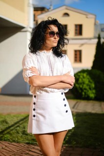 A woman with dark curly hair poses with crossed arms in a fashionable white outfit featuring a