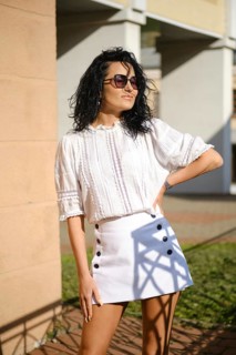 A woman with curly hair stands confidently in a fashionable outfit featuring a white blouse and a