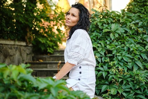 A woman with curly hair is seated gracefully on stone steps surrounded by vibrant green plants. She