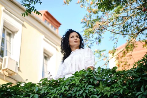 A woman with dark, wavy hair gazes thoughtfully while surrounded by lush green leaves in a charming