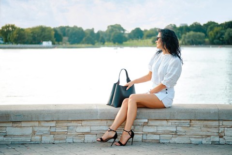 A woman wearing a chic white outfit and black heels relaxes on a stone ledge near calm water. The