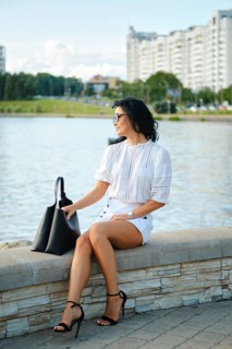 A fashionable woman sits by the water, dressed in a white top and shorts while wearing stylish