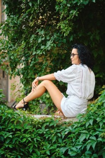 A woman with dark hair sits peacefully in an urban garden surrounded by vibrant green leaves. She
