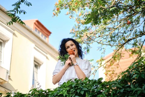 A playful woman stands surrounded by greenery holding a cluster of bright berries. The sun shines