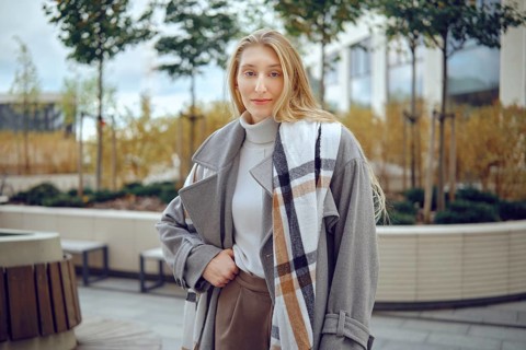 A young woman stands outdoors, showing off her fashionable gray coat featuring a plaid pattern. She