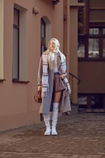 A fashionable young woman in wool coat, knee-high boots and scarf posing in a sunny urban alley.
