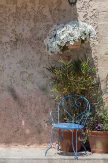 A blue metal chair stands next to potted plants in front of a sand-coloured wall with
