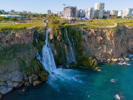 A waterfall cascades from a cliff into the sea, surrounded by urban landscape and green parks,