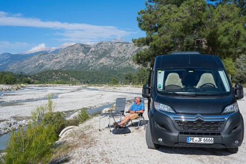 A man sits next to a motorhome on a riverbed surrounded by mountains, camper, Dargaz River near