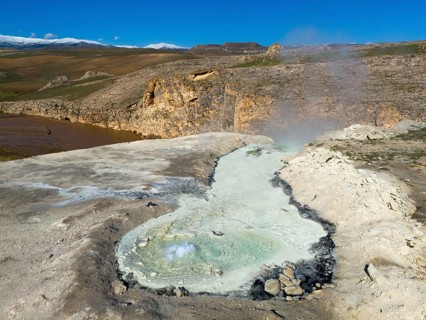 A geothermal area with a steaming geyser and rocks in a wild landscape under a blue sky, aerial