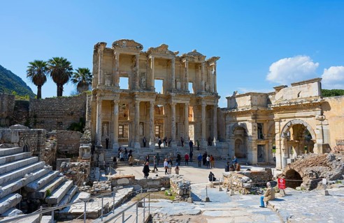 Ancient ruins with imposing columned facades and palm trees, surrounded by visitors under a blue