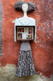 Abstract sculpture of a woman on a rust-red plastered wall. Figure holding a box with photos,