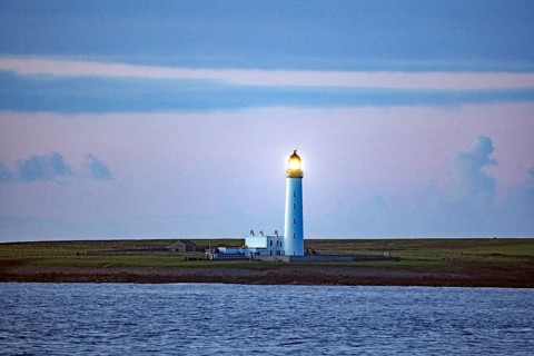 Lighthouse on the coast at dusk with blue sky in the background, crossing between Orkney and