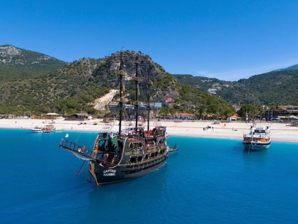 Pirate ship sails in front of a picturesque coastal landscape with mountains and clear blue water,