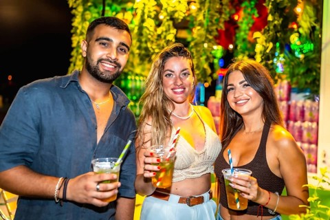 Three friends smiling and holding cocktails at a vibrant outdoor night party, enjoying the festive