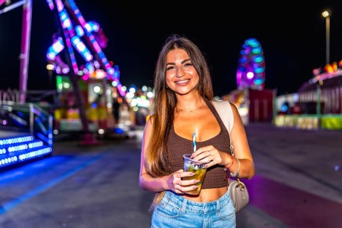 Young woman holding a refreshing drink at a lively amusement park, enjoying the vibrant atmosphere