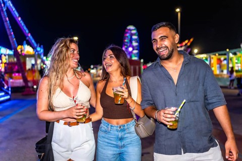 Three friends walking and laughing together at amusement park at night, holding drinks and enjoying