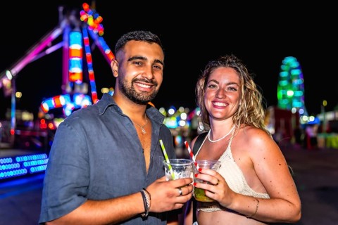 Young man and woman smiling, holding cocktails, enjoying a night out at amusement park with