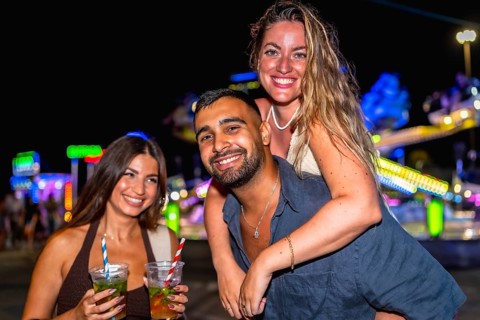 Three friends are having fun at an amusement park at night, enjoying cocktails and the festive