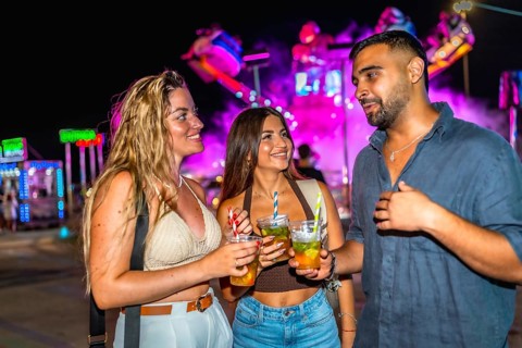 Three friends are holding cocktails and chatting at an amusement park at night, enjoying the