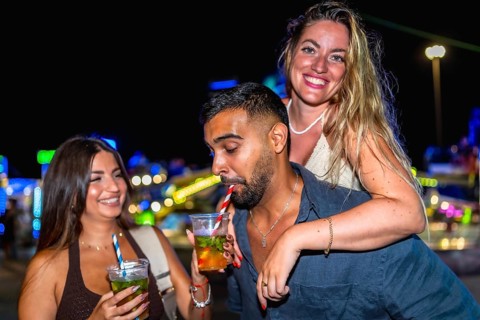 Three friends are having cocktails at an amusement park at night, enjoying the vibrant atmosphere