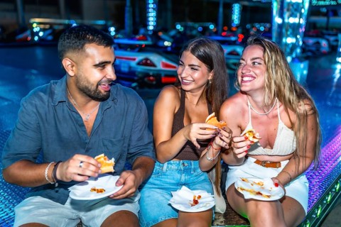 Three happy friends are enjoying burgers and pizza at an amusement park, illuminated by colorful