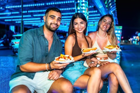 Three friends are sitting and smiling, holding hot dogs on paper plates at a brightly lit amusement