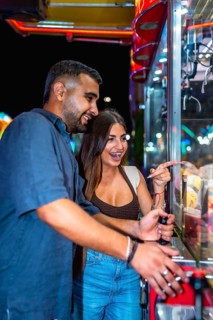 Young couple having fun playing a claw crane game at an amusement park at night, trying to win a