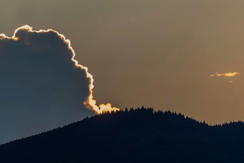 A radiant sunset conjures up warm colours as the clouds hang over a mountain range. Trees line the