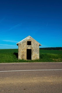 A rustic stone hut stands alone by a quiet road, surrounded by vibrant green fields. Auvergne.