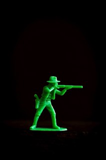 A vibrant green plastic toy cowboy stands in a tactical pose, aiming a rifle against a dark