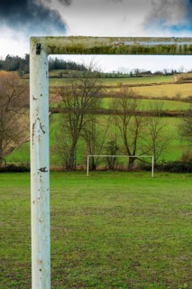 A weathered soccer goalpost stands in the foreground of a wide grassy field, with lush green hills