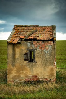 A small, dilapidated hut with a broken roof and cracked walls sits alone in a vast green field.