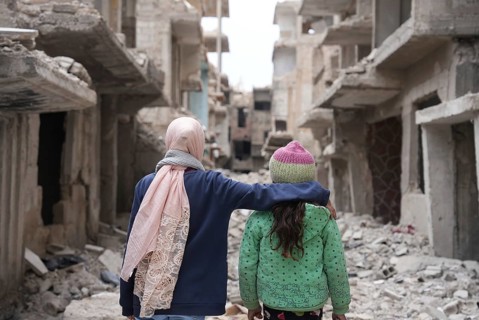Two girls stand in shock in front of a building destroyed by the war in Syria