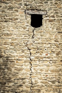 A weathered stone wall displays a prominent crack running vertically, featuring a small window.