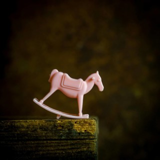 A small pink rocking horse toy sits on a weathered wooden shelf, gently illuminated in low light.