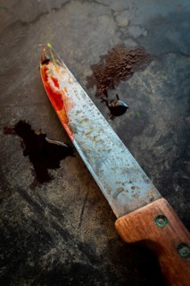 A worn knife with a rusty blade and wooden handle rests on a dark, textured surface. Traces of red
